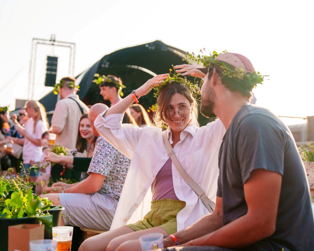 To studerende sidder til Kontur studiestartsfestival og laver blomsterkranse. De har begge en på hvert deres hoved. I baggrunden ses en musikscene og andre studerende