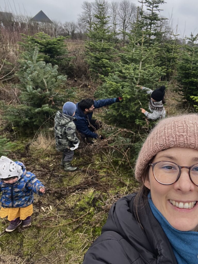 Selfie af Maja med sin mand og to børn i baggrunden, der er ved at fælde et juletræ