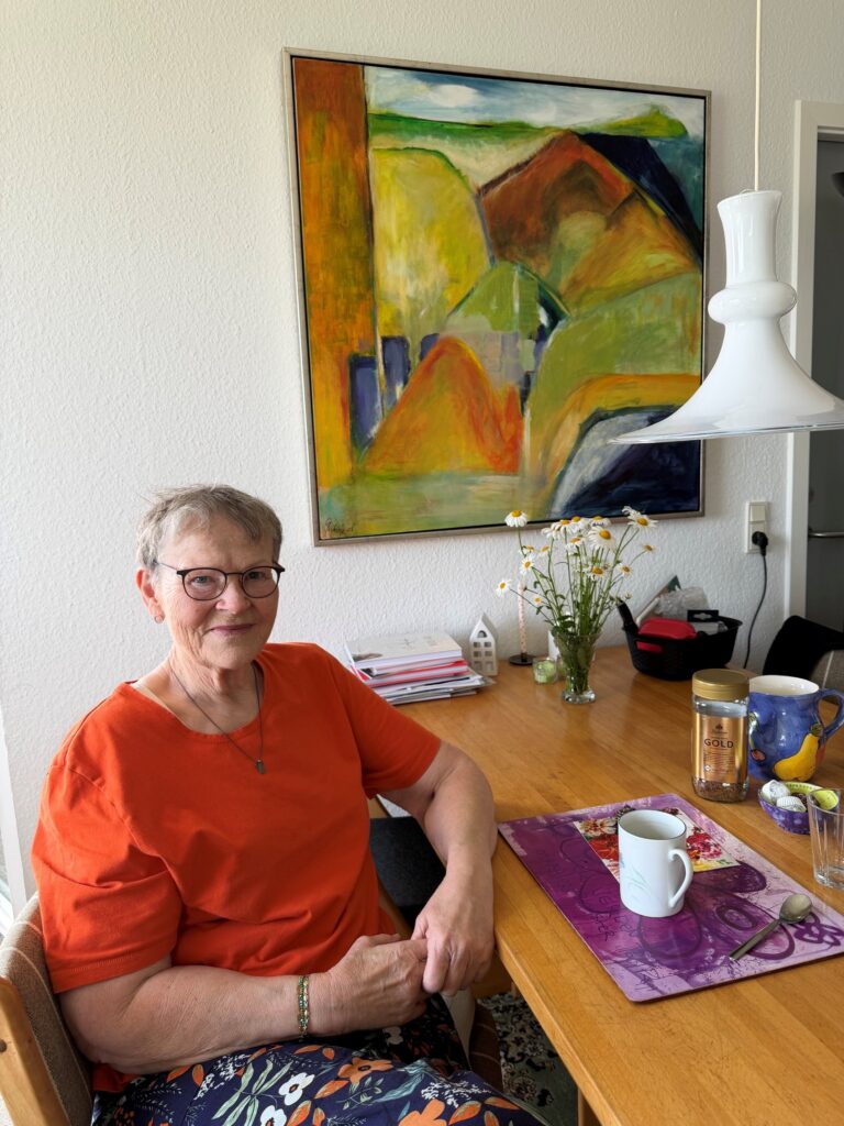 Gerthie Elsen sidder ved sit spisebord. På væggen bag hende hænger et maleri, hun selv har malet