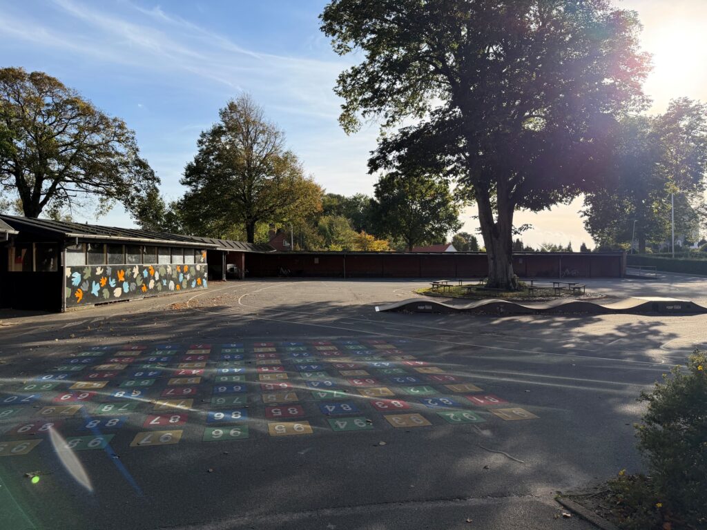 Harte Skoles skolegård med malede felter med tal på asfalten, et stort træ i højre side og et farvestrålende skur i venstre side
